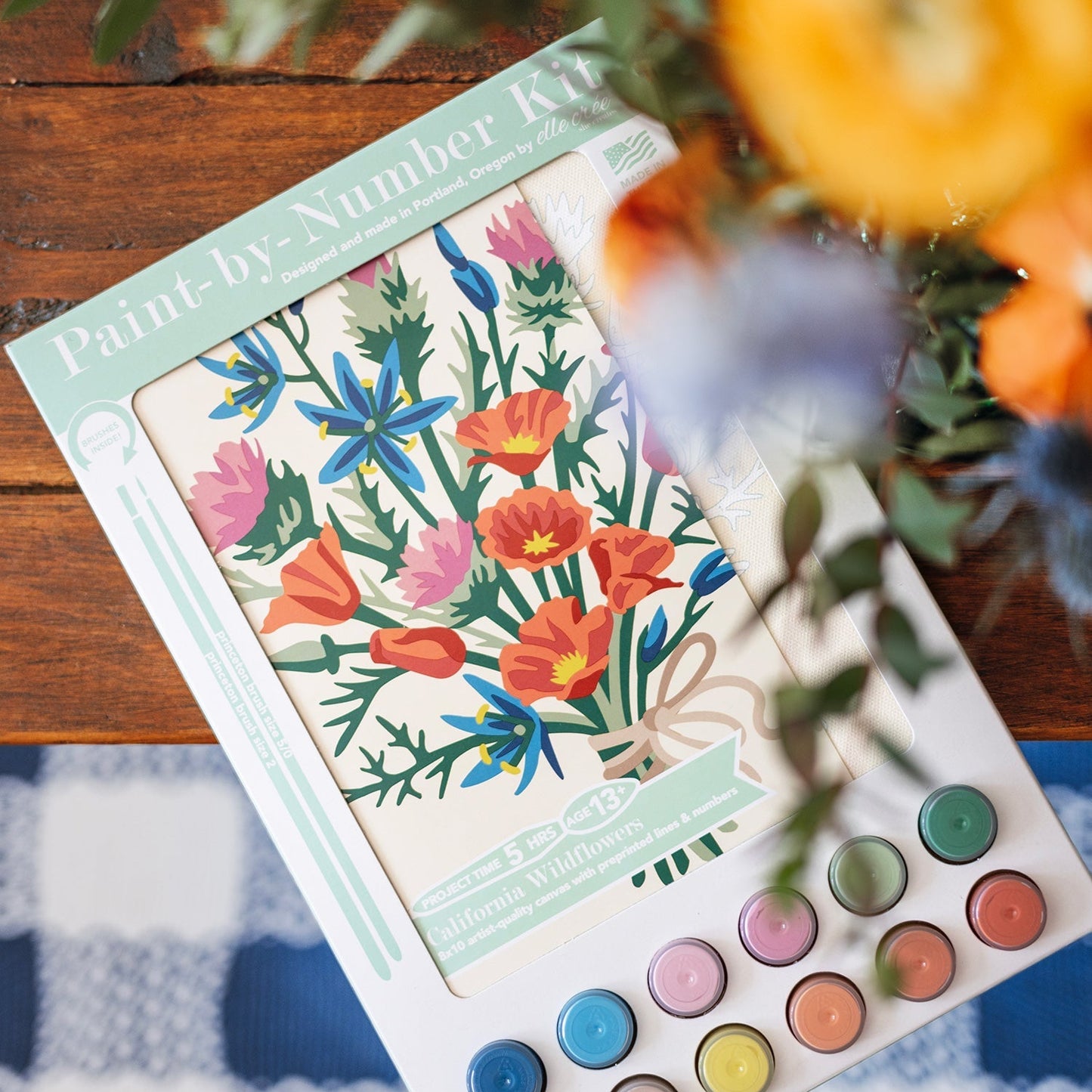 Paint-by-Number kit with floral design and watercolors on a checkered tablecloth.