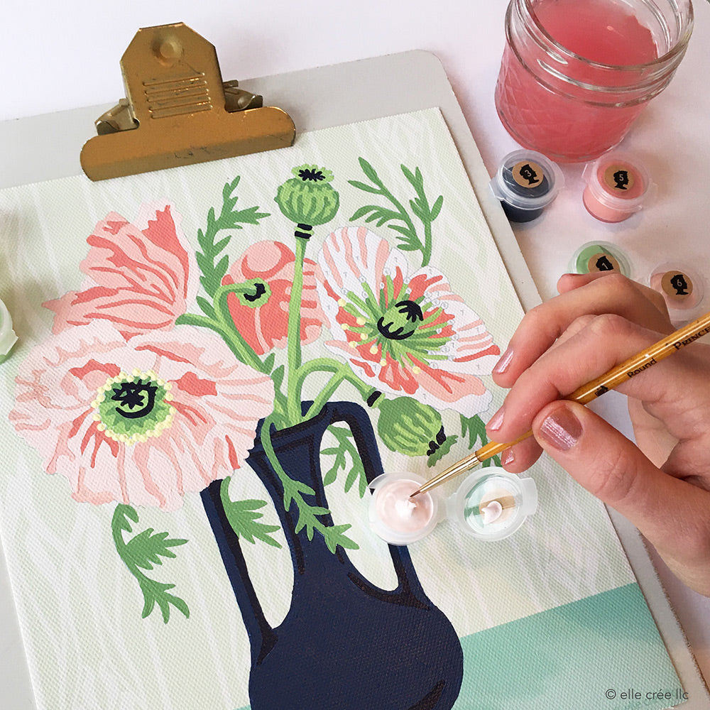 Person painting a floral design on paper with a paintbrush, using a jar of pink paint and a gold clip as props.